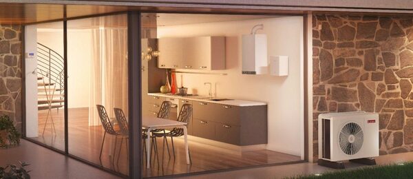 An Ariston air-to-water heat pump outdoor unit installed beside a modern glass-walled house, with a wall-mounted indoor unit visible in the kitchen