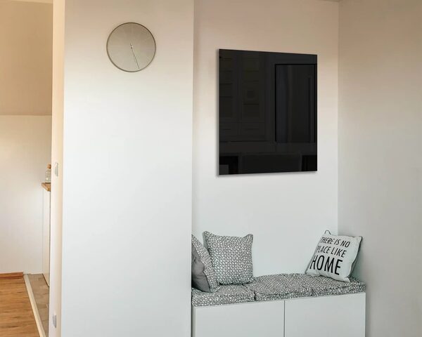 A black glass infrared heating panel mounted on a white wall above a cushioned bench seat in a home hallway
