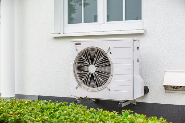 An air source heat pump unit mounted on the exterior wall of a white house below a window