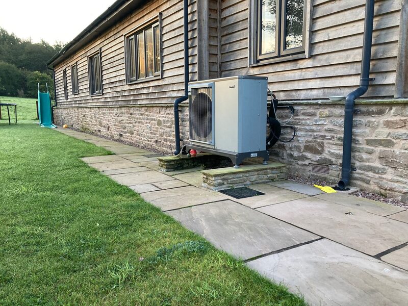 An air source heat pump unit installed on a plinth outside a stone and timber-clad house beside a paved patio