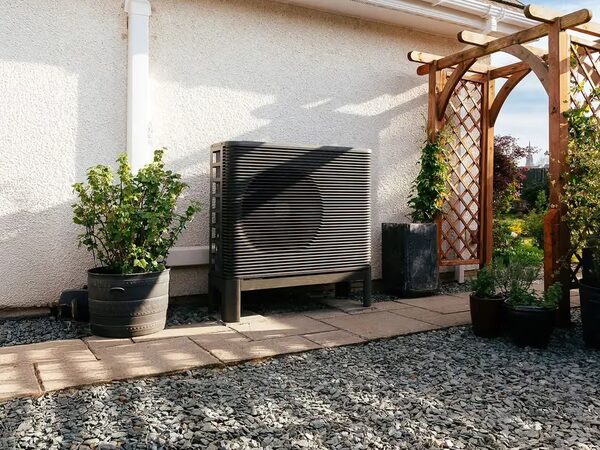 An air source heat pump unit installed outside a house wall beside a garden path and wooden pergola