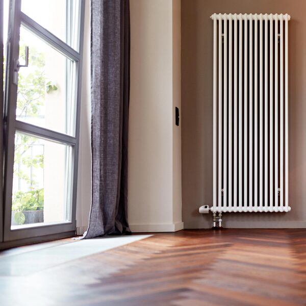 A tall white column radiator with a thermostatic valve on the wall of an empty room beside a large window with a grey curtain and wooden floor