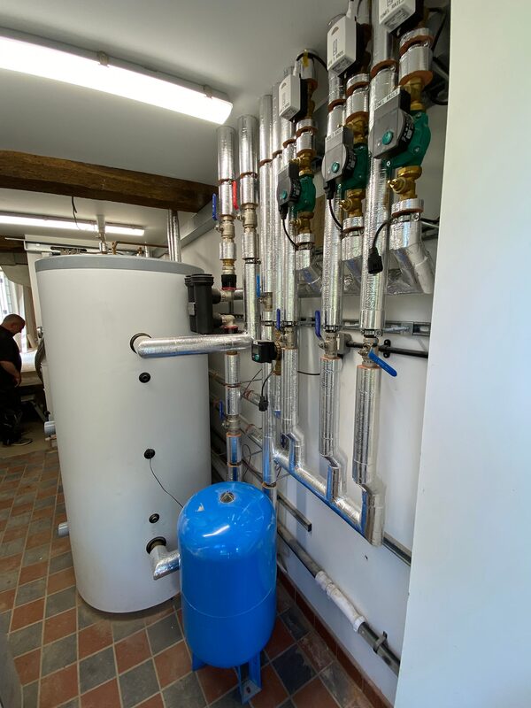 A large white hot water cylinder with insulated pipework, valves and a blue expansion vessel installed in a plant room as part of a ground source heat pump system