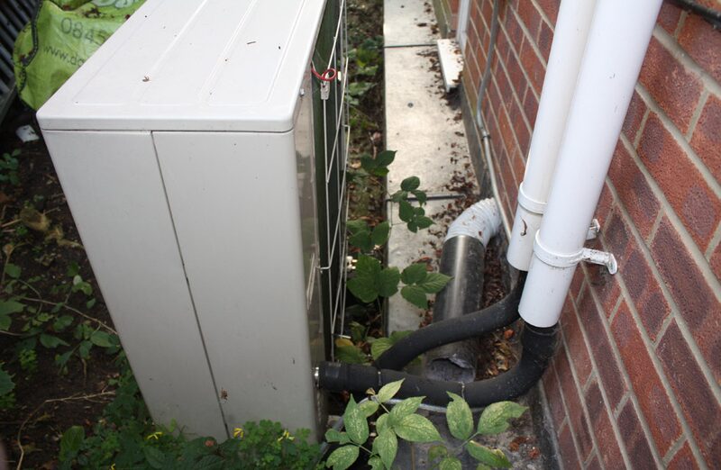 An outdoor air source heat pump unit installed beside a brick house wall with pipework and weeds around it