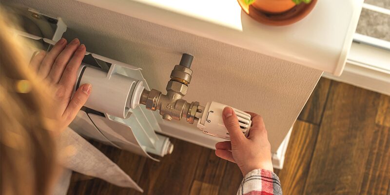 A person turning the thermostatic radiator valve on a white radiator under a windowsill