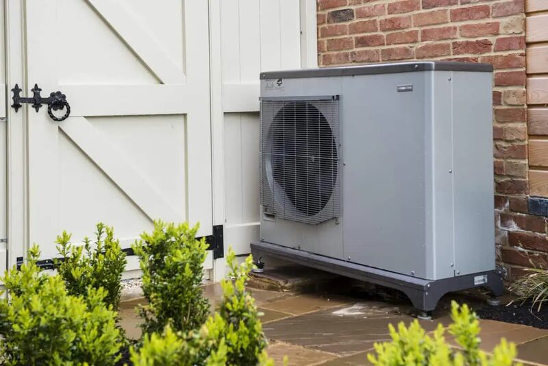 A grey air source heat pump unit installed outside a brick house beside a cream garage door, with shrubs in the foreground