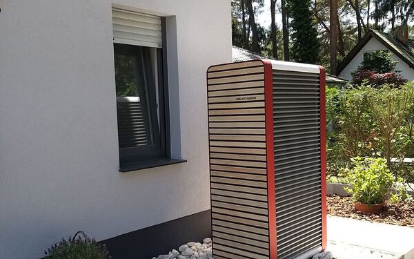 An outdoor air-source heat pump unit with red trim standing on gravel beside the white exterior wall of a house