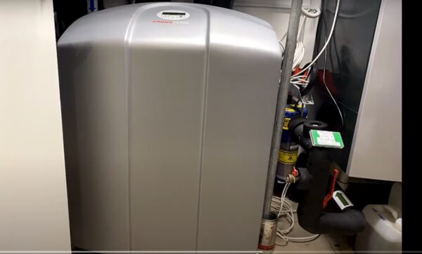 A silver Kronoterm heat pump water heater cylinder installed in a utility cupboard with pipework and valves beside it