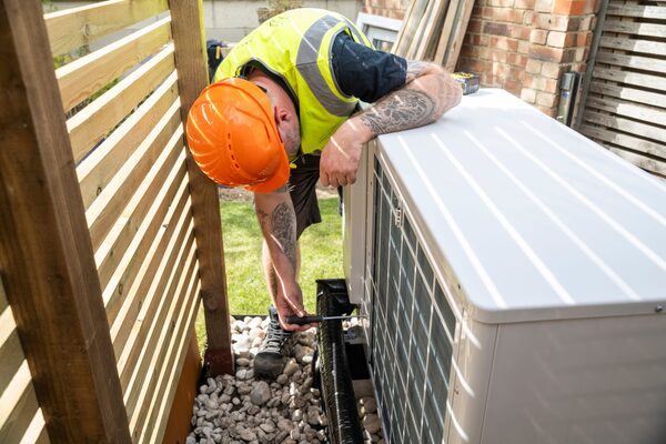 A worker in an orange hard hat and hi-vis vest installing an air source heat pump unit in a garden beside a wooden slatted screen