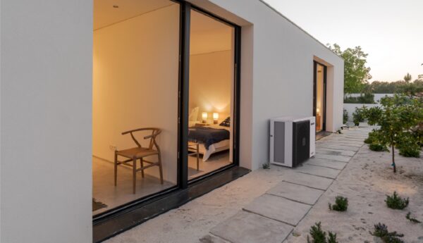 A Hitachi air source heat pump unit installed on the exterior wall of a modern white house at dusk, with lit interior visible through large glass doors