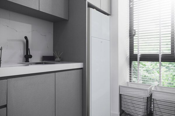 A tall white hot water cylinder installed in a grey utility room next to a sink and window