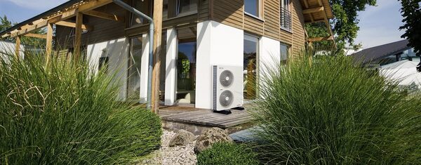 An air source heat pump unit installed on a wooden deck outside a modern timber-clad house, surrounded by tall grasses