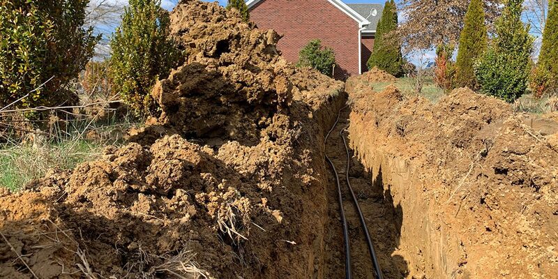 A deep trench dug in a garden with black ground-loop pipework laid inside, for a ground source heat pump installation outside a brick house