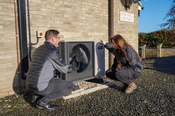 An engineer and a woman crouch beside an Ideal air source heat pump installed outside a brick cottage