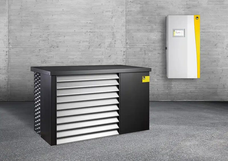 An iDM AERO heat pump outdoor unit with louvered grille beside a wall-mounted white and yellow control unit against a concrete wall