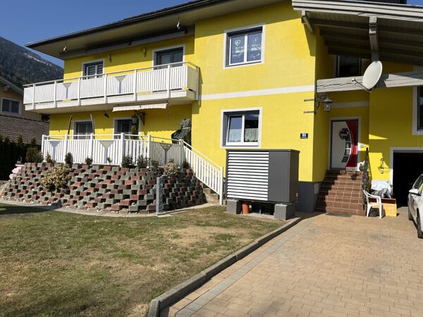 A grey heat pump outdoor unit installed beside a yellow two-storey house with a driveway