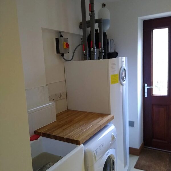 A tall white IVT ground source heat pump cylinder installed in a utility room next to a washing machine, with pipework and an isolation switch above