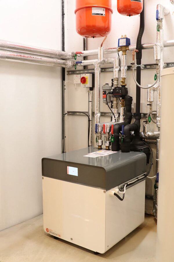 A Kensa ground source heat pump unit installed in a plant room with pipework, valves and orange expansion vessels