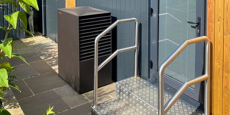 A dark grey outdoor heat pump unit installed against the exterior wall of a building beside a door with metal handrails and steps