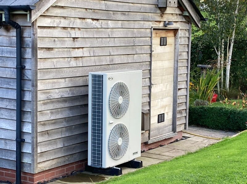 An air source heat pump unit installed outside a weatherboard-clad garden building on a lawn