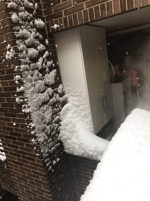 An outdoor heat pump unit recessed into a brick wall, heavily covered in snow during snowfall