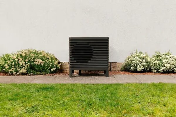 A dark grey Aira air source heat pump unit installed outside a house against a white wall, with shrubs and a lawn in front