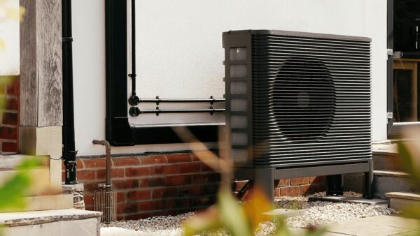 A dark grey air source heat pump unit installed outside a house next to a white rendered wall and brick base