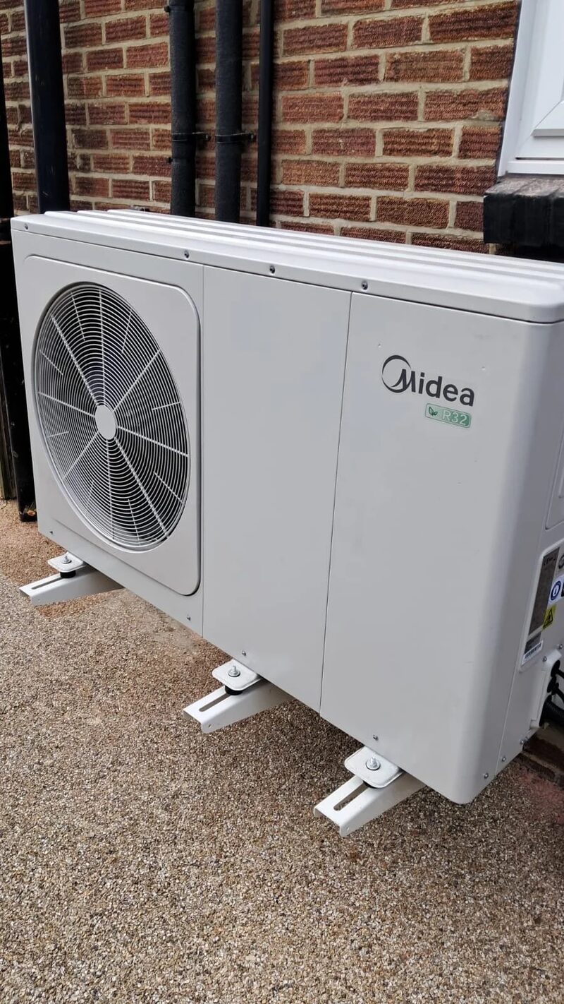 A white Midea R32 air source heat pump unit installed on mounting brackets outside a brick house