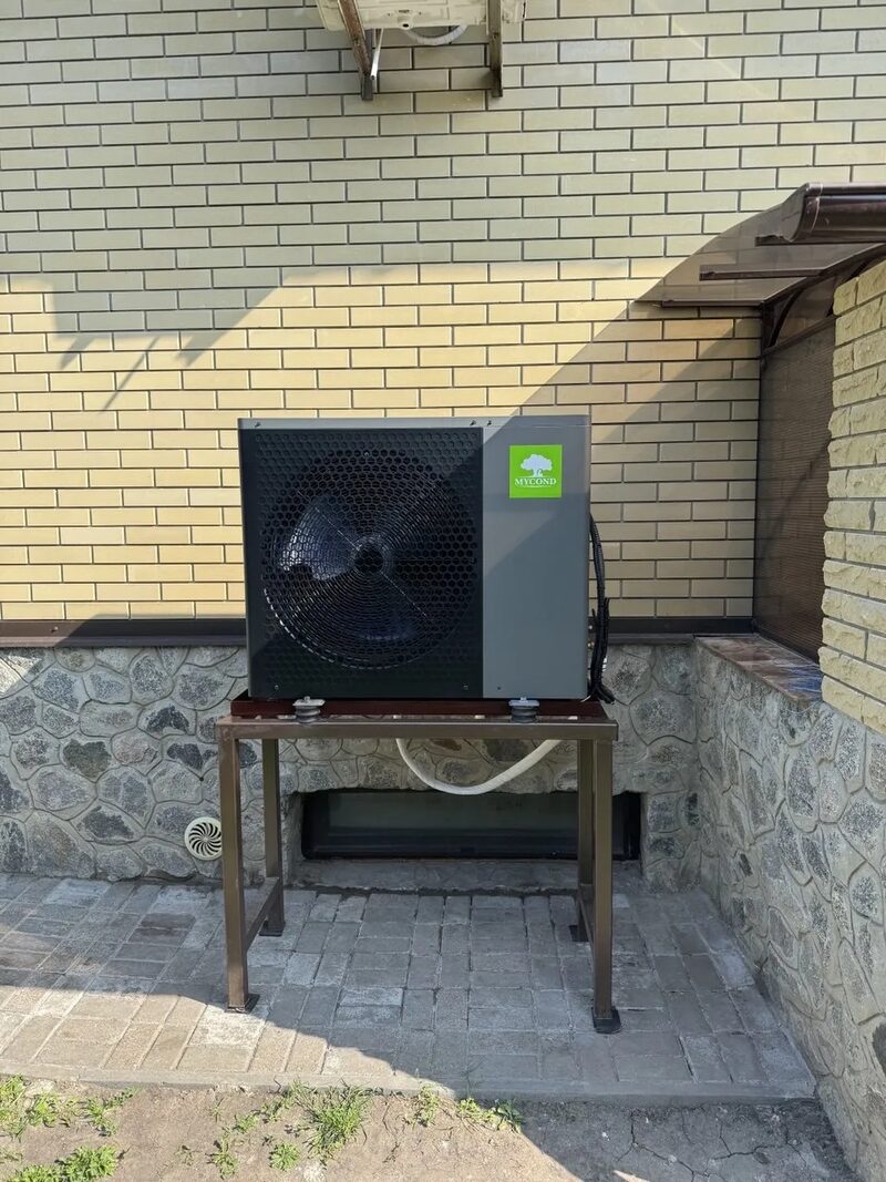 A Mycond BeeHeat split heat pump outdoor unit installed on a metal stand against the brick wall of a house