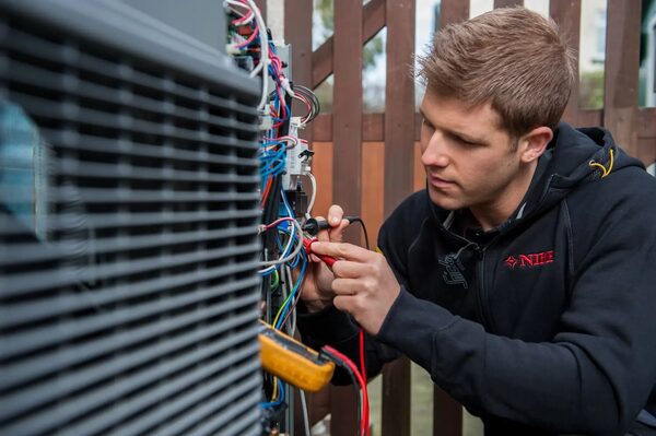 A technician in a NIBE-branded hoodie using a multimeter to test wiring on an outdoor heat pump unit