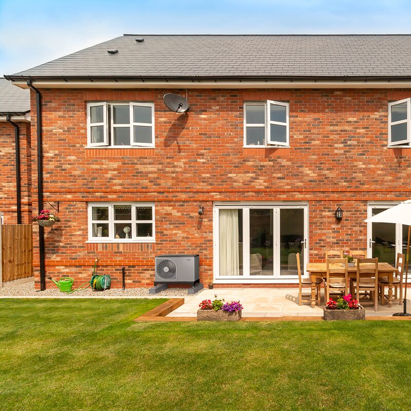 A NIBE air source heat pump unit installed on gravel against the back wall of a brick house with a patio and garden