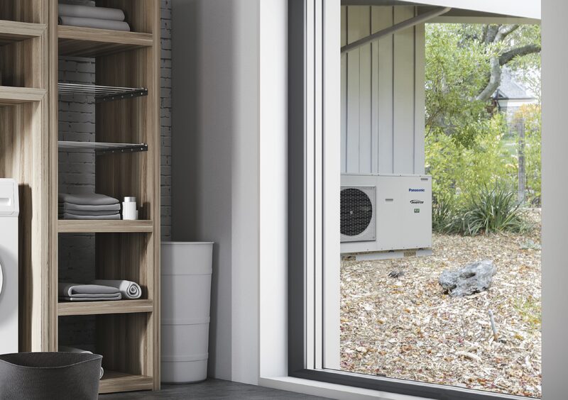 A Panasonic Aquarea air-to-water heat pump monobloc unit installed outside a house, seen through a window from a utility room