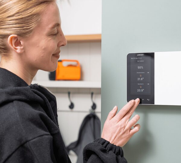 A woman adjusting a wall-mounted Passiv smart thermostat showing hot water and heating temperatures