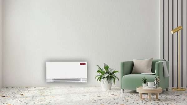 A white Heatboost fan-assisted radiator mounted low on a wall in a styled living room with a green armchair and plant