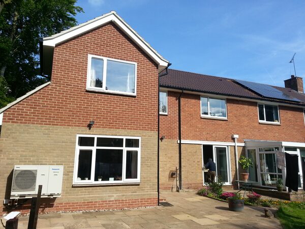 A Panasonic air source heat pump unit mounted on the outside wall of a brick house with solar panels on the roof