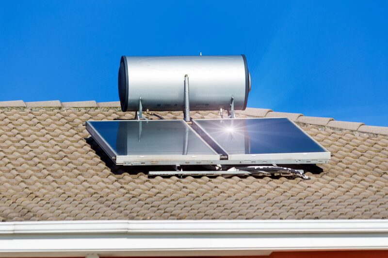 A rooftop solar thermal water heating system with two flat-plate collector panels and a cylindrical hot water storage tank mounted on a tiled roof against a blue sky