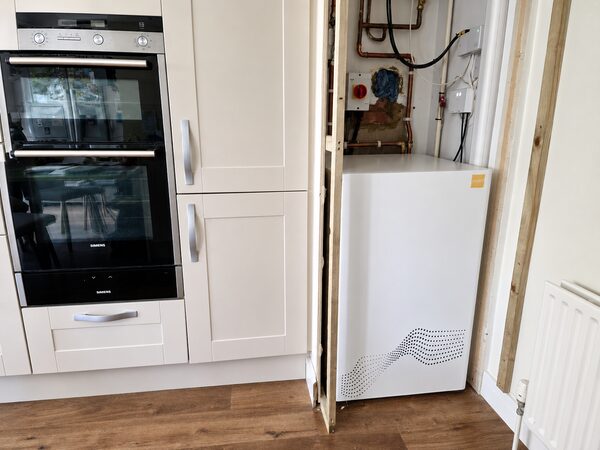 A white tepeo heat battery boiler installed in a kitchen cupboard next to a Siemens double oven, with exposed pipework and wiring above