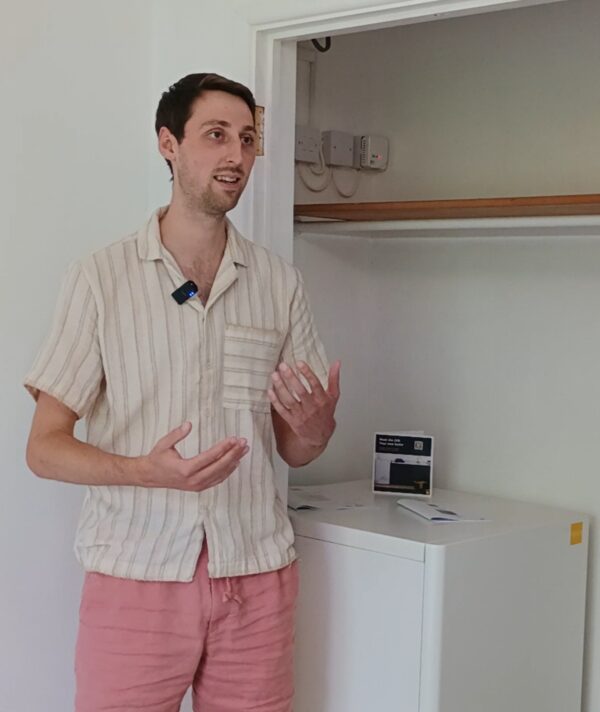A man gesturing beside a white tepeo ZEB electric heating battery installed in a flat, with a leaflet on top