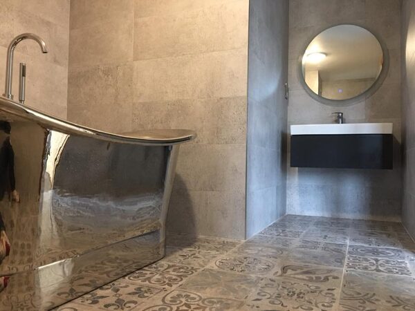 A tiled bathroom with a freestanding metal bath, wall-mounted basin and round mirror, showing patterned tiled floor suitable for underfloor heating