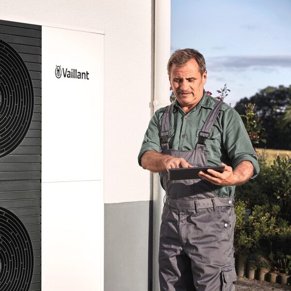 A technician in overalls using a tablet next to an outdoor Vaillant air source heat pump unit mounted on a house wall