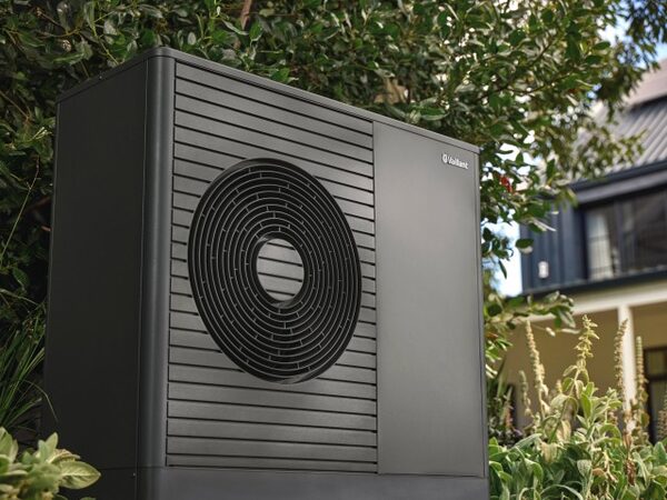A dark grey Vaillant aroTHERM plus air source heat pump unit installed outside a house among garden plants.