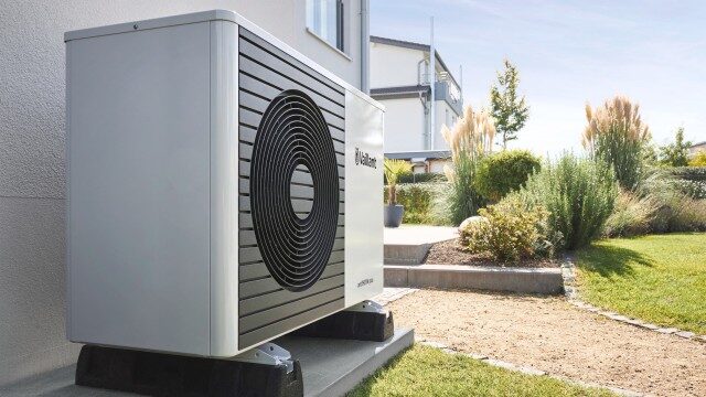 A Vaillant aroTHERM plus air source heat pump unit installed on a plinth outside a house wall beside a garden