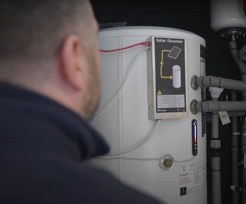 A technician looking at a white Mixergy hot water cylinder fitted with a Solar Diverter unit and pipework in a dark airing cupboard