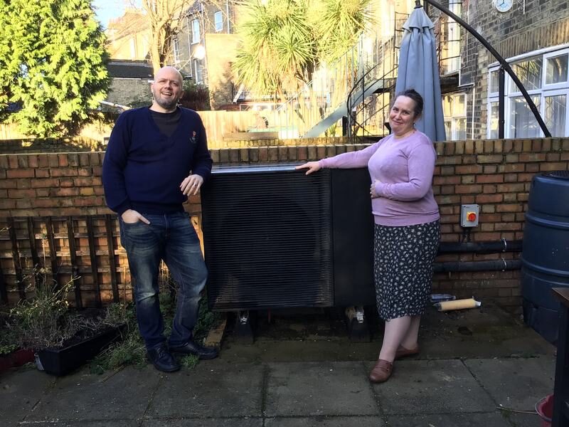 A man and a woman standing in a garden patio beside a large black air source heat pump unit against a brick wall
