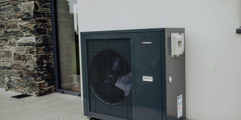 A Warmflow air source heat pump unit installed outside a house wall next to a stone wall and glass door