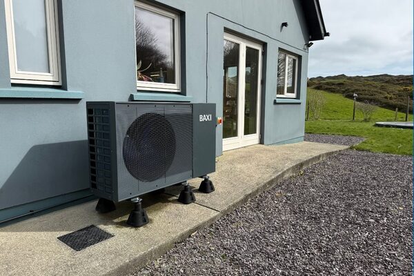 A grey Baxi air source heat pump unit installed on the ground outside a blue-grey house