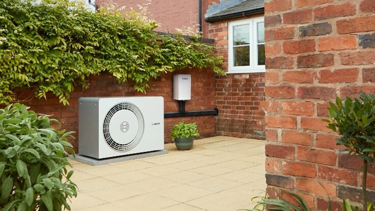 A Worcester Bosch air source heat pump outdoor unit installed on a paved patio beside a brick house wall, with a small wall-mounted unit above it