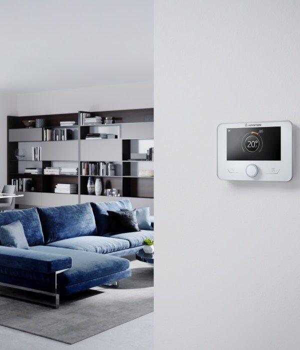 An Ariston Sensys Net wall-mounted smart thermostat displaying 20 degrees on a white wall beside a modern living room with a blue sofa