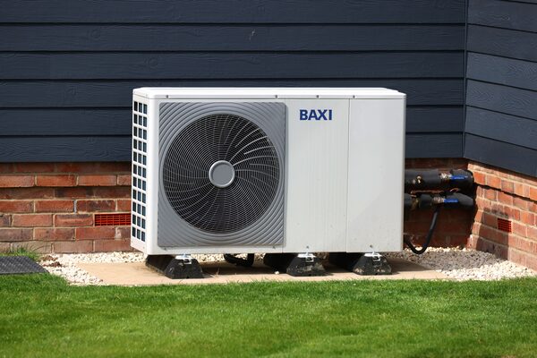 A white Baxi air source heat pump unit installed outside a house against a dark grey clad wall with brick base and gravel