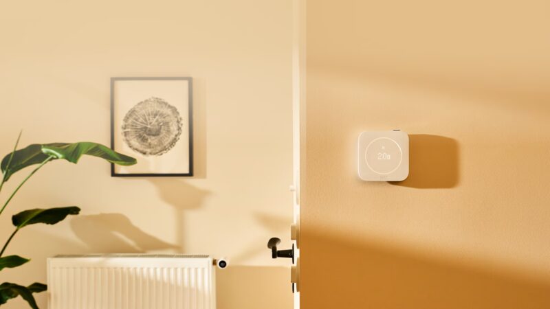A tado° Smart Thermostat X mounted on a warm-toned wall above a radiator in a sunlit room